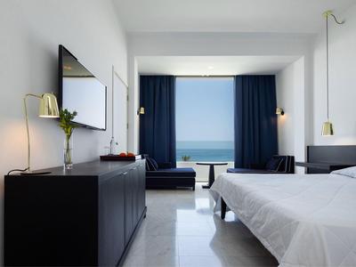 Helles Hotelzimmer mit Doppelbett, dunklen Möbeln und Blick auf das Meer durch große Fenster.