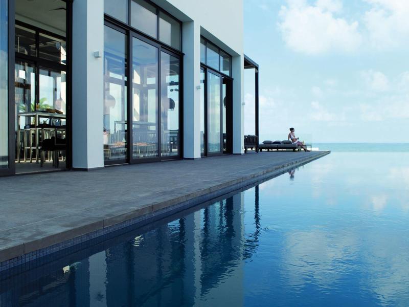 Moderner Pool mit dunklem Holzdeck, Glasfassade und ruhigem blauem Wasser unter blauem Himmel.