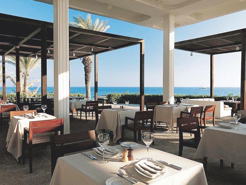 Offenes Restaurant mit weißen Tischen, Stühlen und Meerblick im Hintergrund bei Tageslicht.