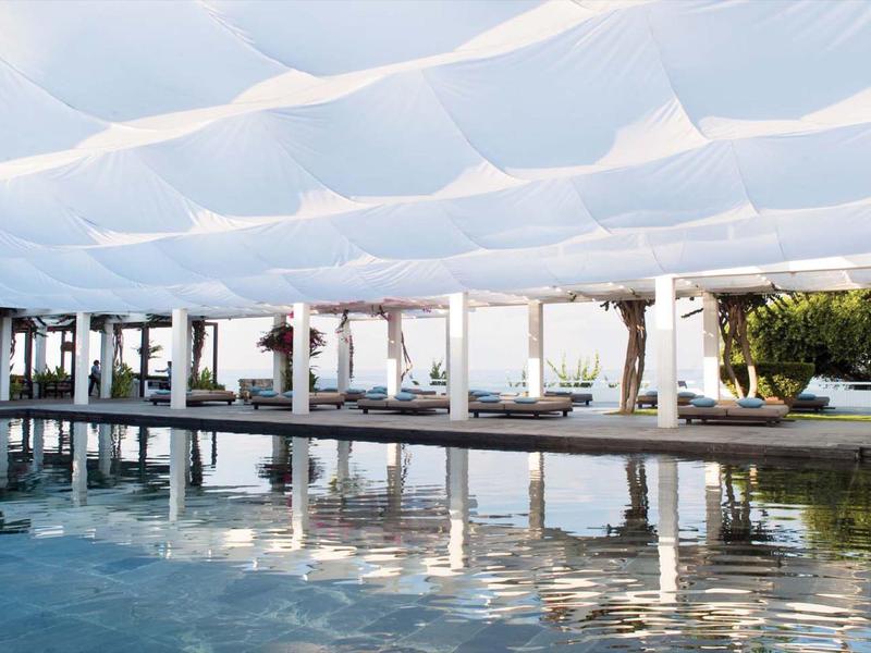 Moderner Außenpool mit blauem Wasser, darüber weiße, gewellte Sonnensegel, umgeben von Säulen.