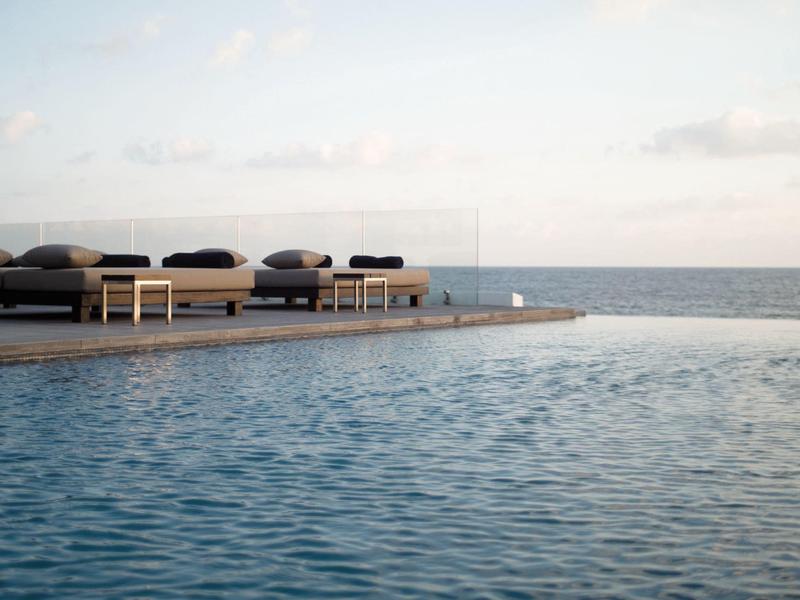 Infinity-Pool mit blauer Wasseroberfläche und Liegen am Rand, dahinter Meer und Himmel.