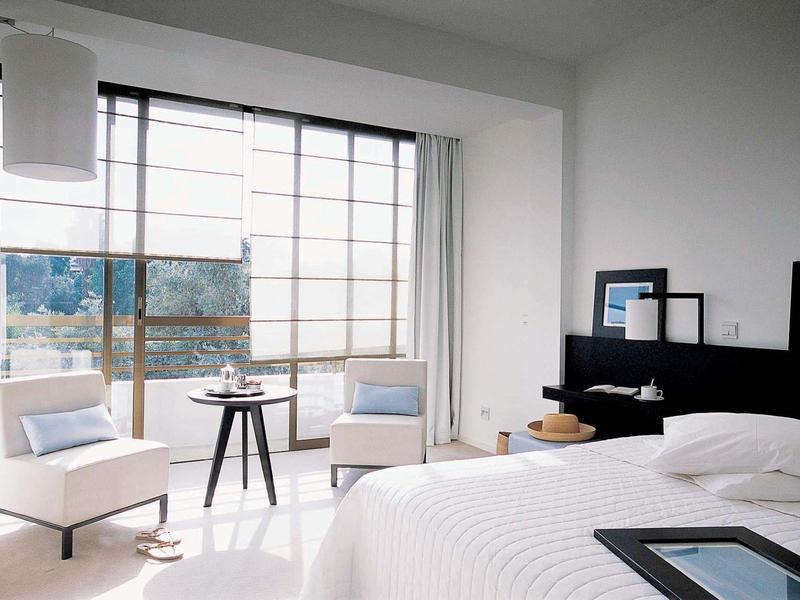 Hell eingerichtetes Hotelzimmer mit großem Bett, zwei Sesseln und bodentiefen Fenstern.