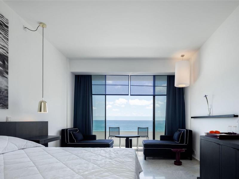 Moderner weißer Raum mit großem Fenster, Meerblick, Bett, dunklen Möbeln und Vorhängen.