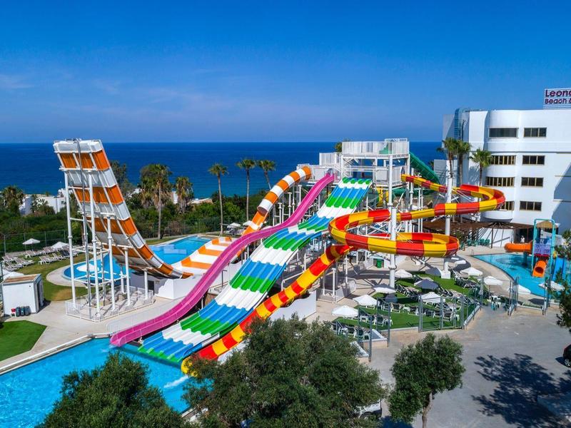 Bunte Wasserrutschen führen in einen Pool, umgeben von Bäumen und einem weißen Hotelgebäude.