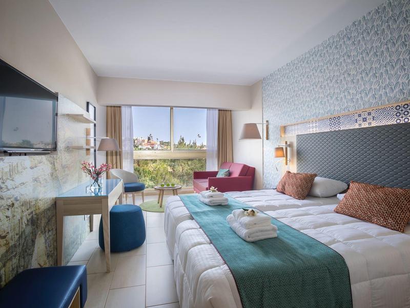 Modernes Hotelzimmer mit Doppelbett, Glasfront und Sitzbereich mit rotem Sofa und Balkonblick.