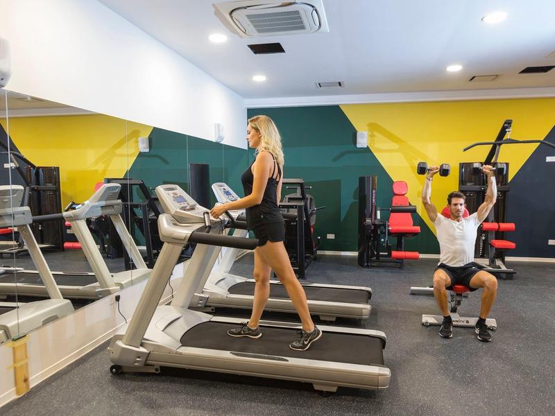 Frau läuft auf Laufband, Mann trainiert mit Kabelzug in hellem, buntem Fitnessraum.