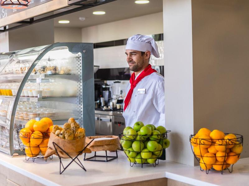 Ein Mann in Kochkleidung steht hinter einer Theke mit Körben voller grüner und gelber Früchte.