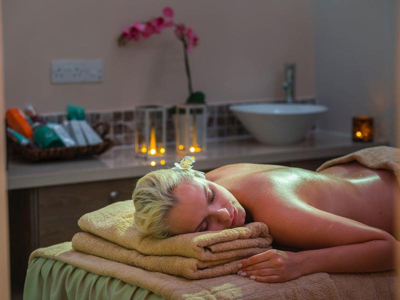 Entspannte Frau liegt auf Massagetisch, umgeben von Handtüchern, Kerzenlicht und Orchidee.