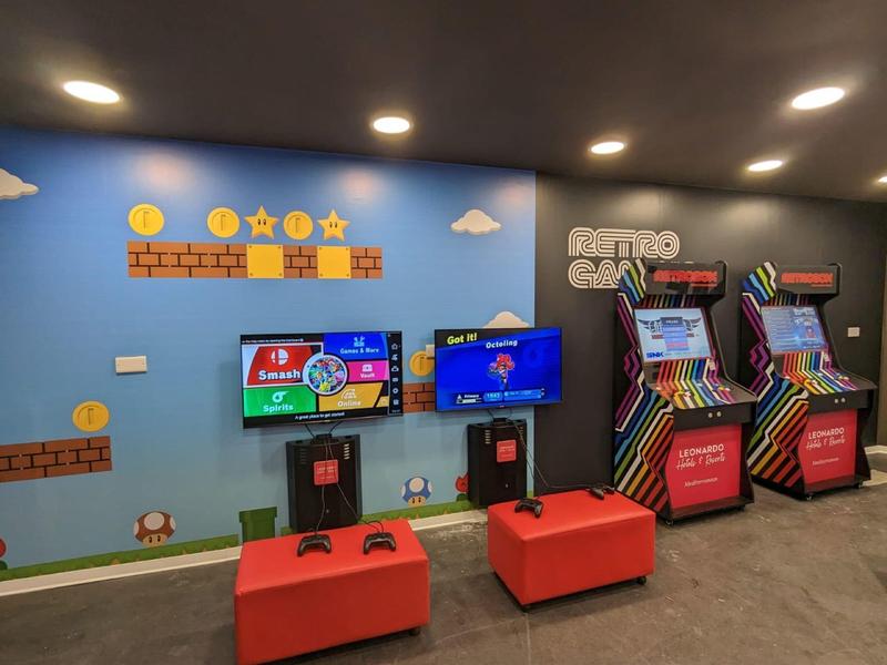 Ein Gaming-Bereich mit blauer Wand, Retro-Spielautomaten und zwei modernen Konsolen mit roten Sitzhockern.