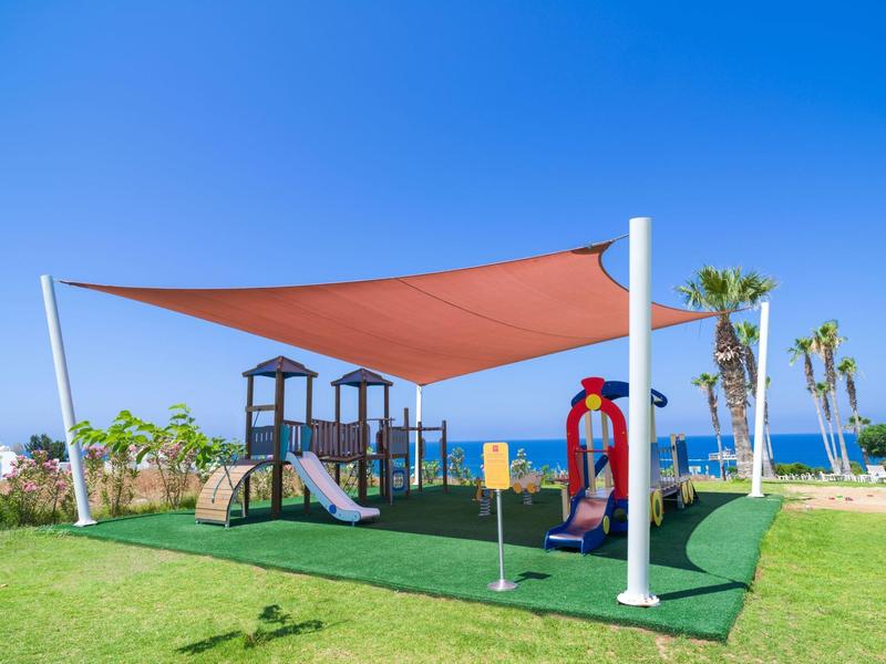 Spielplatz mit zwei Rutschen, rotem Sonnensegel und künstlichem Grün vor blauem Himmel am Meer.