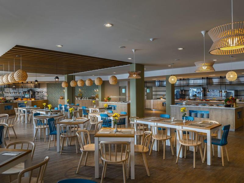 Heller, moderner Gastraum mit Holztischen, bunten Stühlen und runden Lampen an der Decke.