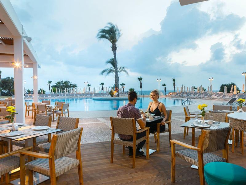 Terrasse mit Tischen und Stühlen am Pool, Menschen sitzen, Palmen und bewölkter Himmel im Hintergrund.