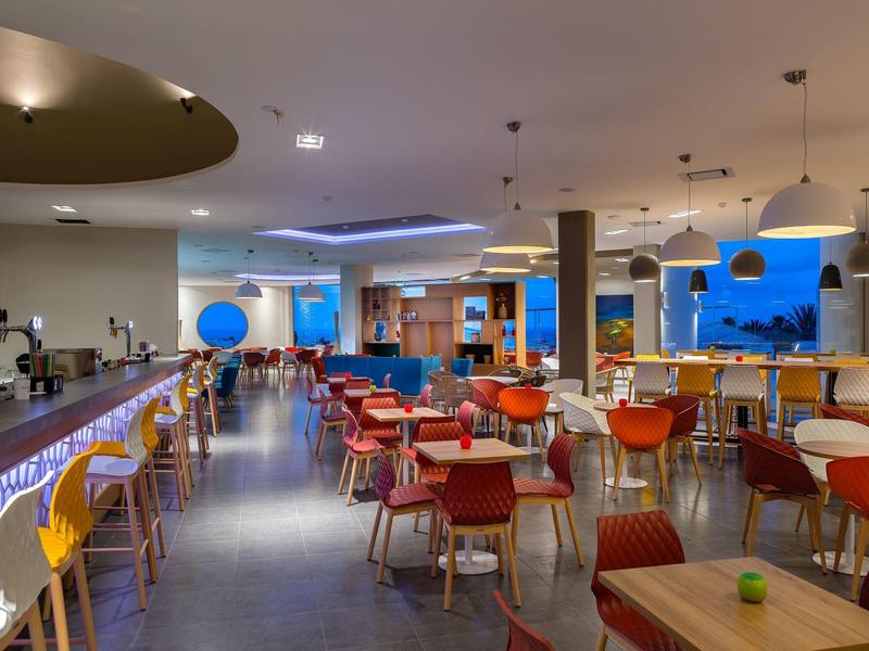 Modernes Café mit bunten Stühlen, Holztischen und großem Aquarium an der Wand.