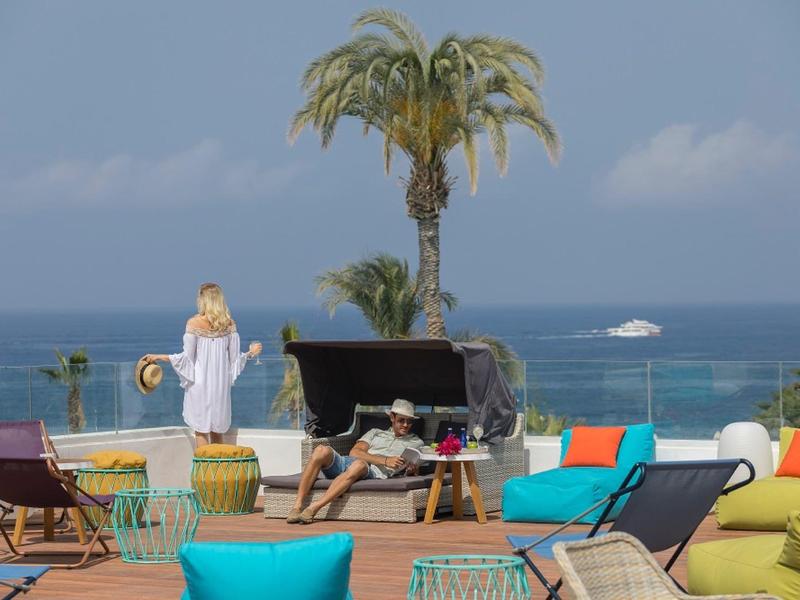 Dachterrasse mit bunten Sitzmöbeln, Palme in großem Topf und Meer im Hintergrund bei Sonnenschein.