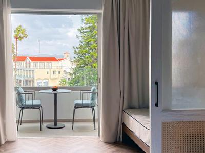 Camera d'hotel con balcone, due sedie e piccolo tavolo con vista su edifici e alberi.