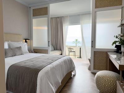 Habitación de hotel luminosa con cama grande, zona de estar y vistas al mar a través de ventanas de suelo a techo.