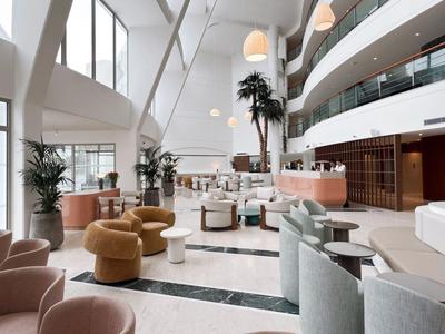 Lobby de hotel luminosa con muebles modernos, grandes ventanas y espacios abiertos de varios niveles.