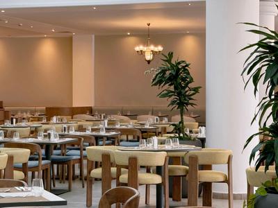 Comedor moderno de hotel con mesas puestas, sillas de madera y plantas grandes.
