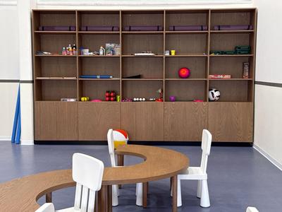 Área de salón de hotel moderna con mesa de madera semicircular, sillas blancas y estantería decorativa.