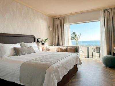 Habitación de hotel luminosa con cama doble, zona de estar acogedora y vista al mar por grandes ventanas.