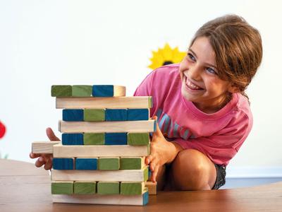 Ein lachendes Mädchen spielt mit einem gestapelten Holzblockturm auf einem Tisch.