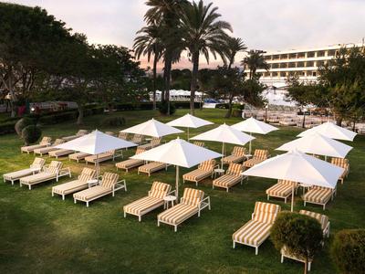 Hotelgarten mit Liegestühlen und weißen Sonnenschirmen auf grünem Rasen unter Palmen.