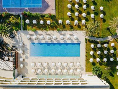 Vogelperspektive eines Hotels mit Pool, Sonnenschirmen und Liegestühlen auf grünem Rasen.