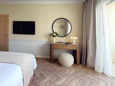 Camera d'hotel luminosa con letto, pavimento in legno, tavolo da trucco, specchio rotondo e tende alla finestra.