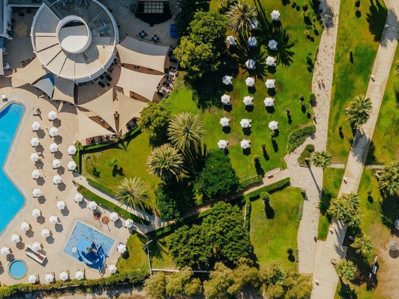 Vogelperspektive auf Hotelpool, Sonnenschirme, Grünflächen und Strand am Meer.