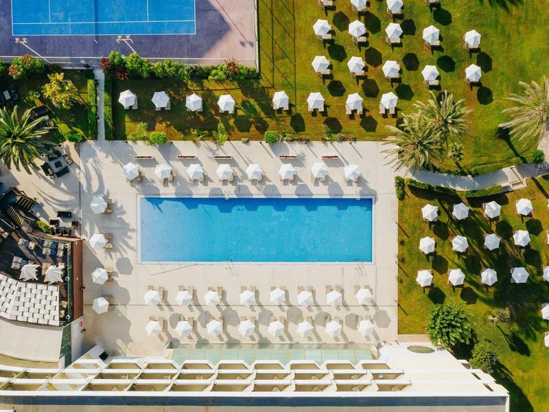 Vogelperspektive eines Hotels mit Pool, Sonnenschirmen und Liegestühlen auf grünem Rasen.