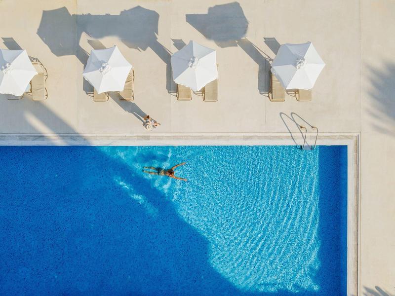 Person schwimmt in klarem Poolwasser neben weißen Sonnenschirmen auf hellem Pooldeck.