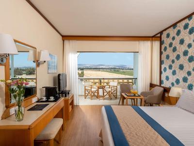 Camera d'hotel luminosa con letto matrimoniale, vista mare e balcone con sedie e tavolo.