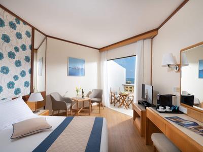 Camera d'hotel luminosa con letto matrimoniale, area salotto, scrivania e balcone con vista mare.