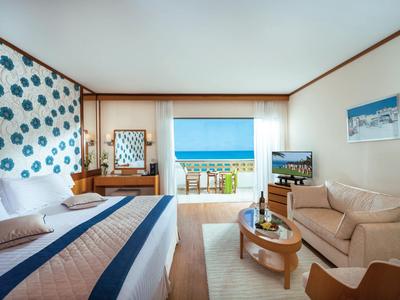 Camera d'hotel luminosa con letto matrimoniale, zona salotto e vista mare attraverso una porta aperta.