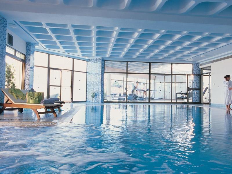 Innenraum eines Schwimmbads mit blauem Wasser, großen Glasfenstern und zwei Personen am Rand.