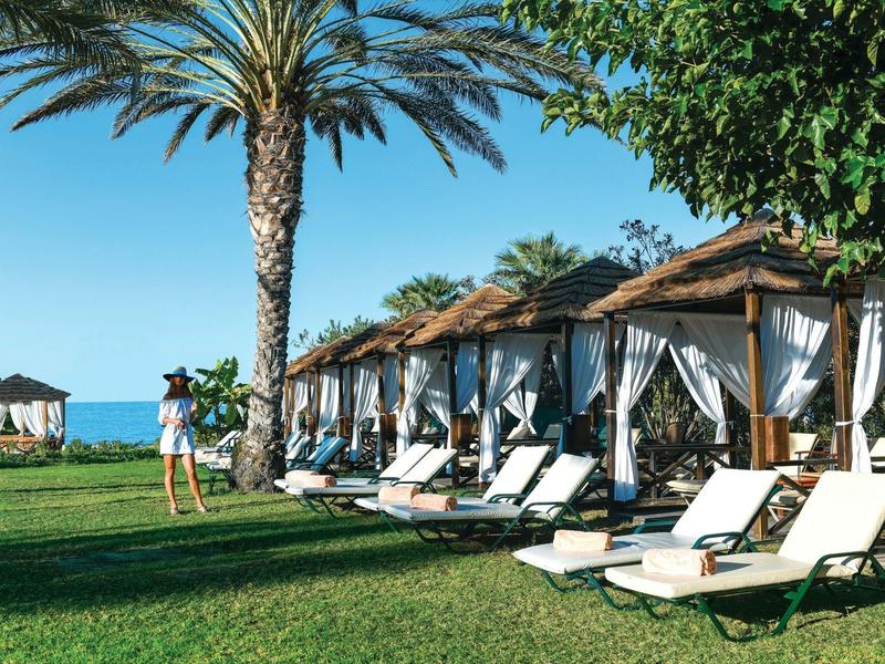 Grüne Wiese mit Palmen, weißen Liegen und bemalten Holzhütten am Meer unter blauem Himmel.