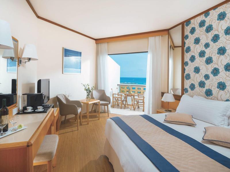 Helles Hotelzimmer mit Doppelbett, Holzmöbeln, Balkon und Meerblick unter blauem Himmel.