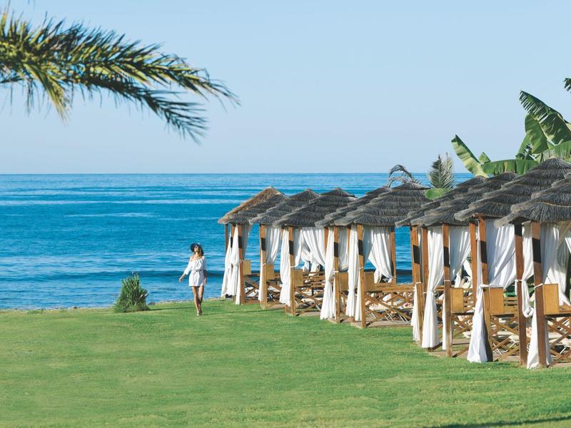 Entspannende Strandliegen mit weißen Vorhängen und Palmen vor dem blauen Meer.