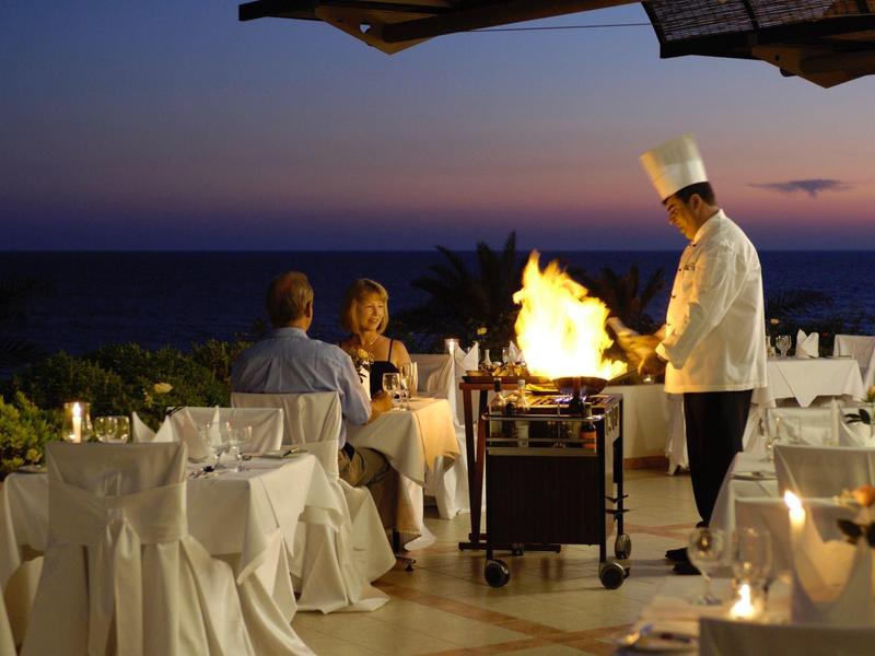 Abendessen auf Terrasse am Meer mit Kellner, der Essen am Grill flambiert, Gäste sitzen am Tisch.
