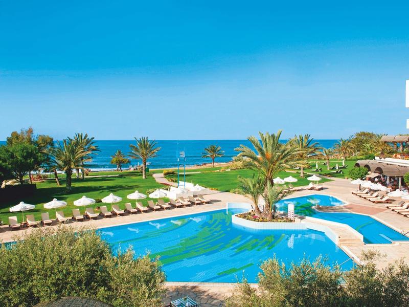 Moderner Pool mit Palmen und Sonnenschirmen, Meer im Hintergrund unter klarem blauem Himmel.