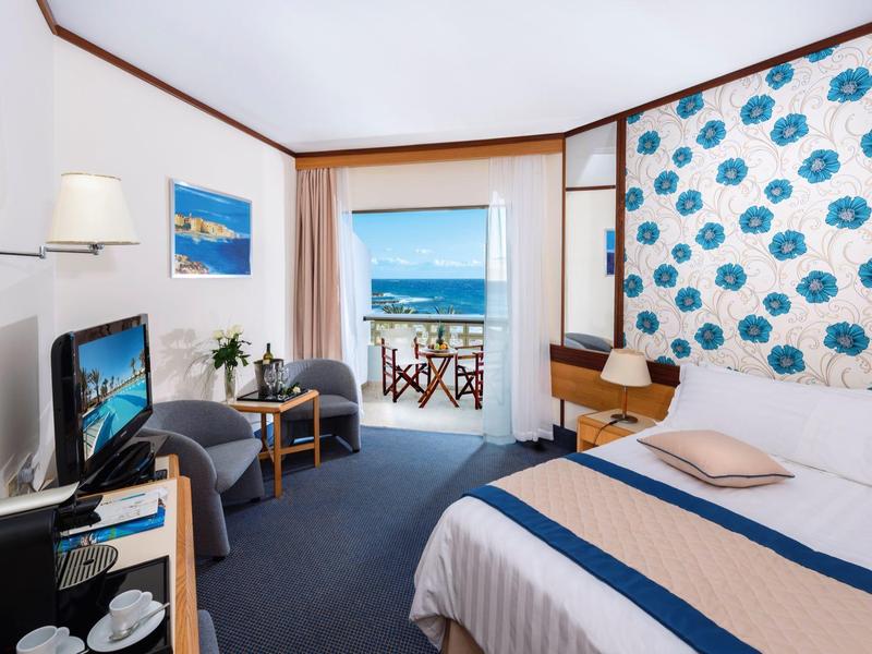 Helles Hotelzimmer mit Meerblick, Doppelbett, Schreibtisch, Sessel und Balkon mit Holzterrasse.