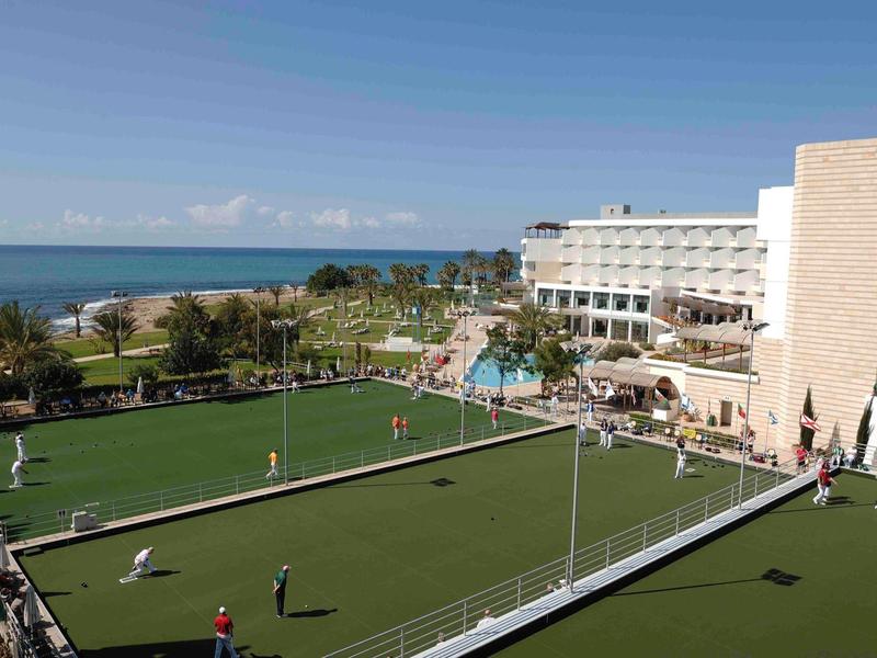Hotelanlage am Meer mit grünem Sportfeld, Pool, Palmen und blauem Himmel.