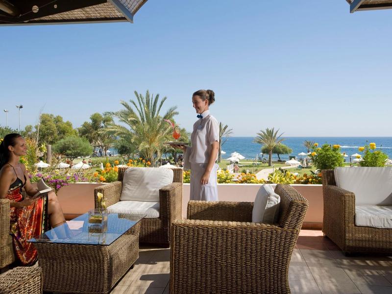 Terrasse mit Korbmöbeln, zwei Frauen, Pflanzen und Meerblick bei klarem Himmel.