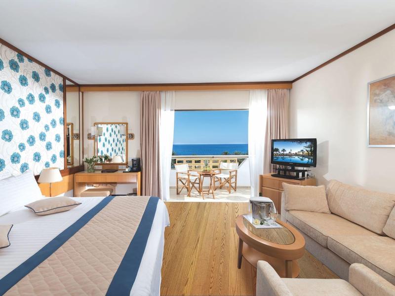 Helles Hotelzimmer mit Doppelbett, Sofa, Fernseher und Balkon mit Meerblick.