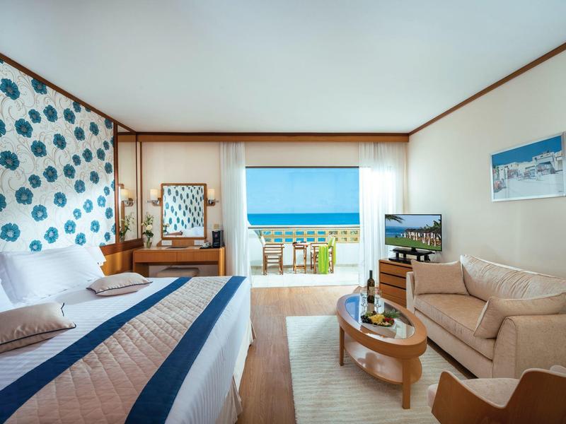 Helles Hotelzimmer mit Doppelbett, blauer Wanddeko, Sofa, Holzmöbeln und Meerblick durch Balkonfenster.