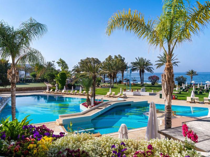 Swimmingpools mit blauer Wasserfläche, Palmen und Liegestühlen am Meer unter klarem Himmel.