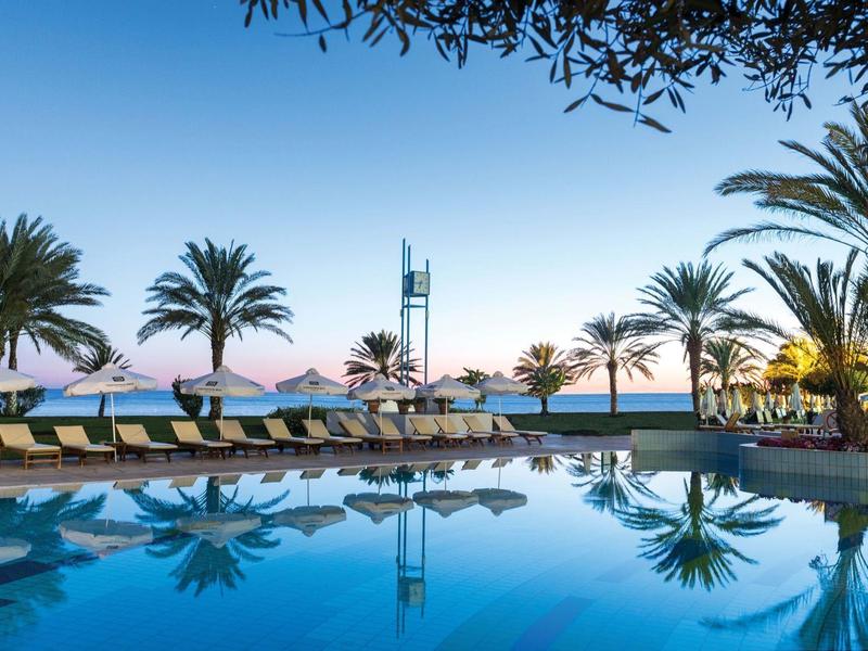 Pool mit Liegen und Palmen vor klarem Himmel und ruhigem Wasser, das Spiegelungen zeigt.