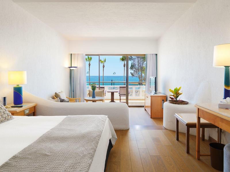 Helles Hotelzimmer mit zwei Betten, Holzboden, Sitzbereich und Meerblick durch große Fenster.