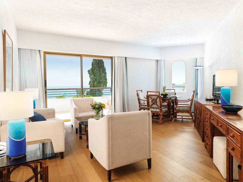 Helles Wohnzimmer mit Sesseln, Holztisch, Sideboard, großen Fenstern und Blick auf Meer und Baum.