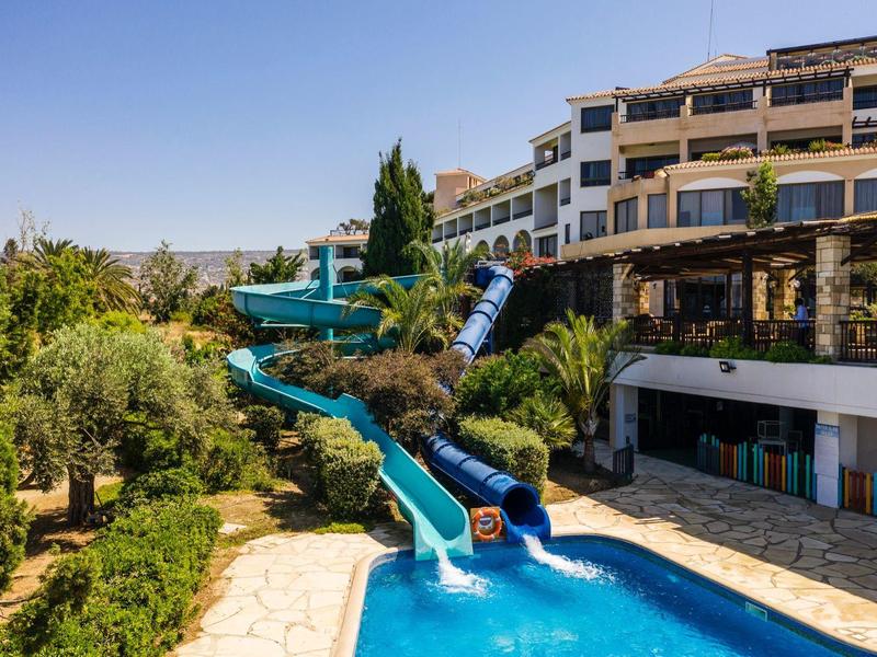 Wasserrutsche führt in blaues Becken vor mehrstöckigem Hotel mit grüner Vegetation bei klarem Himmel.